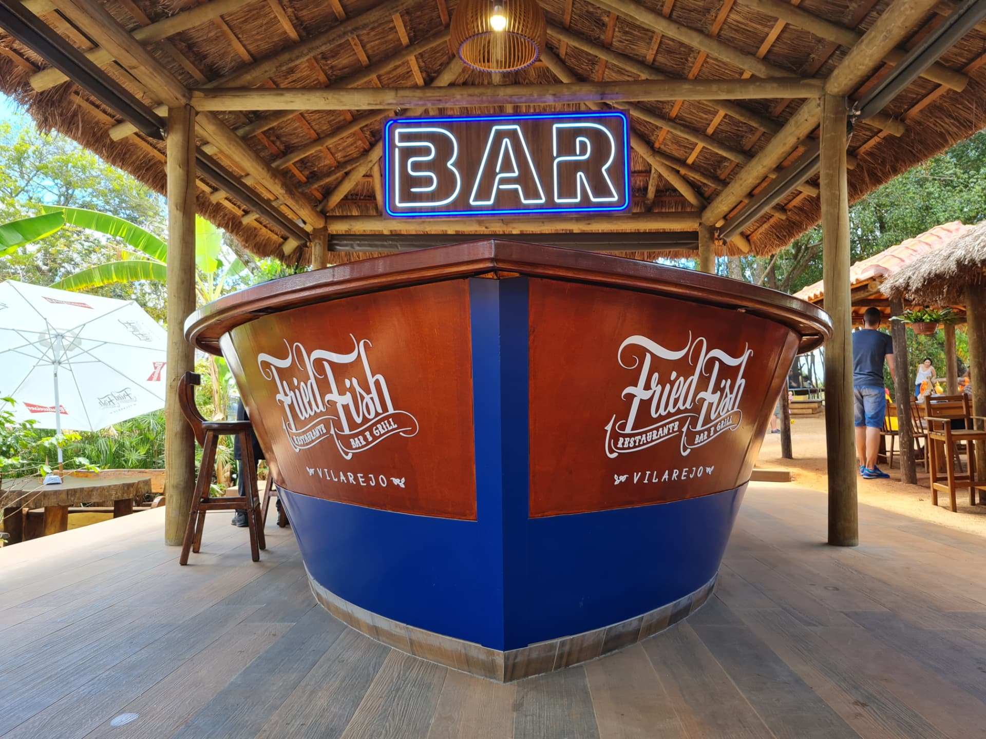 Bar em formato de barco com casco azul e letreiro BAR sob quiosque de palha