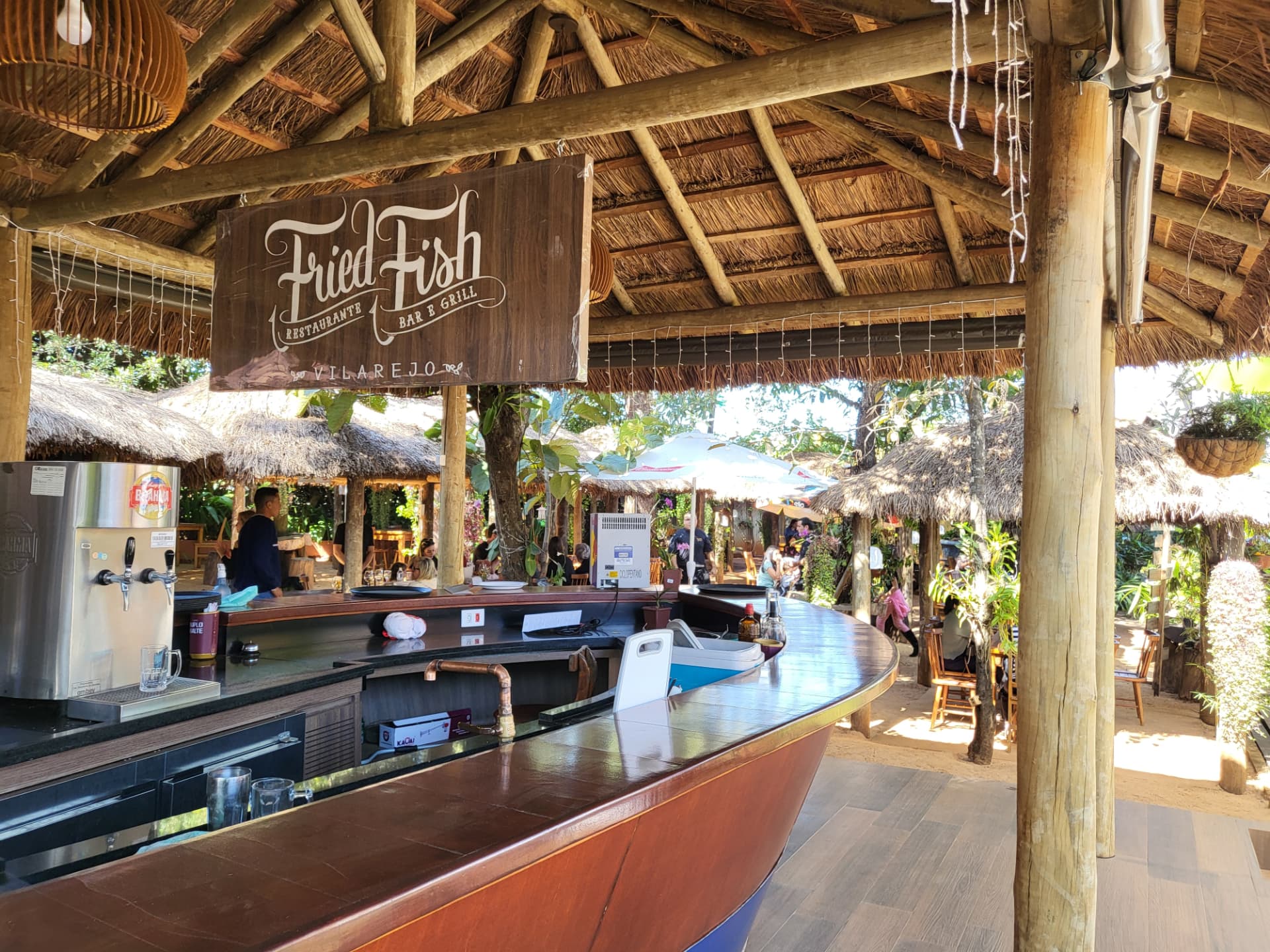 Bar-barco em dia movimentado, com quiosques de palha e vegetação ao fundo