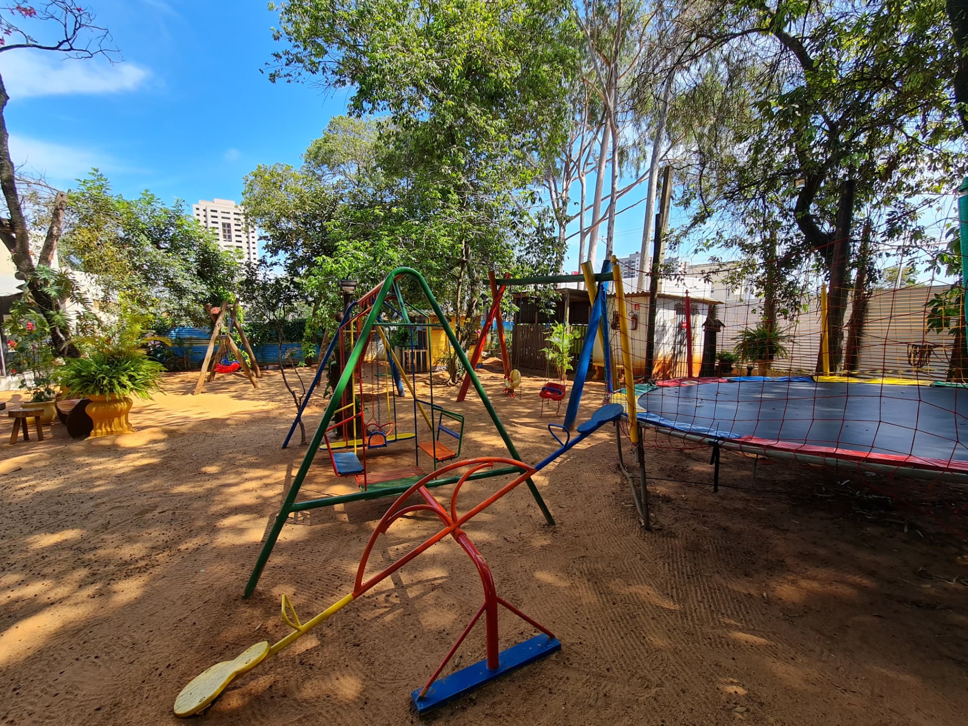 Balanços coloridos, gangorra e cama elástica em área de areia sombreada