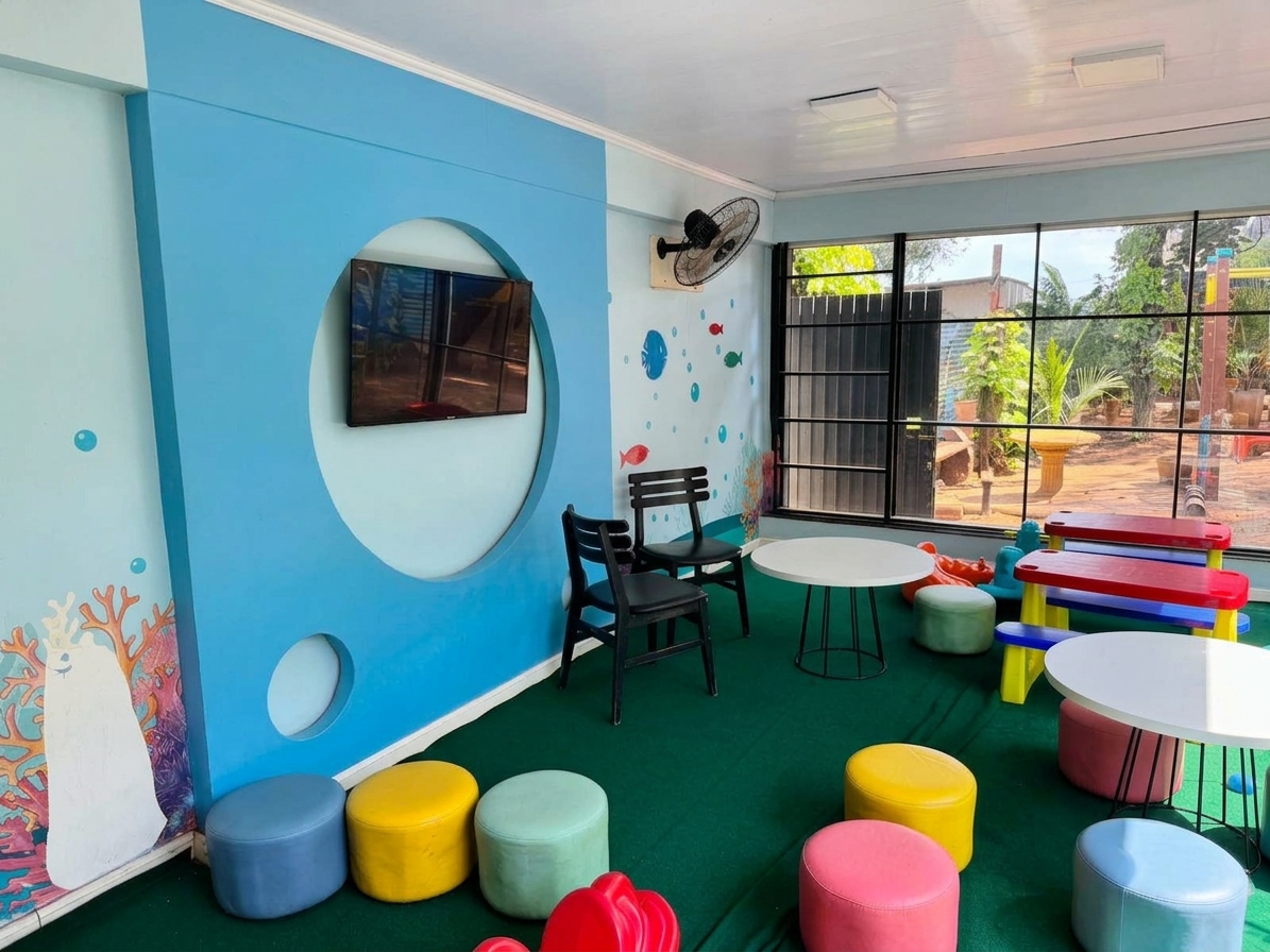Interior da brinquedoteca com TV na parede azul, pufes coloridos e mesa de piquenique