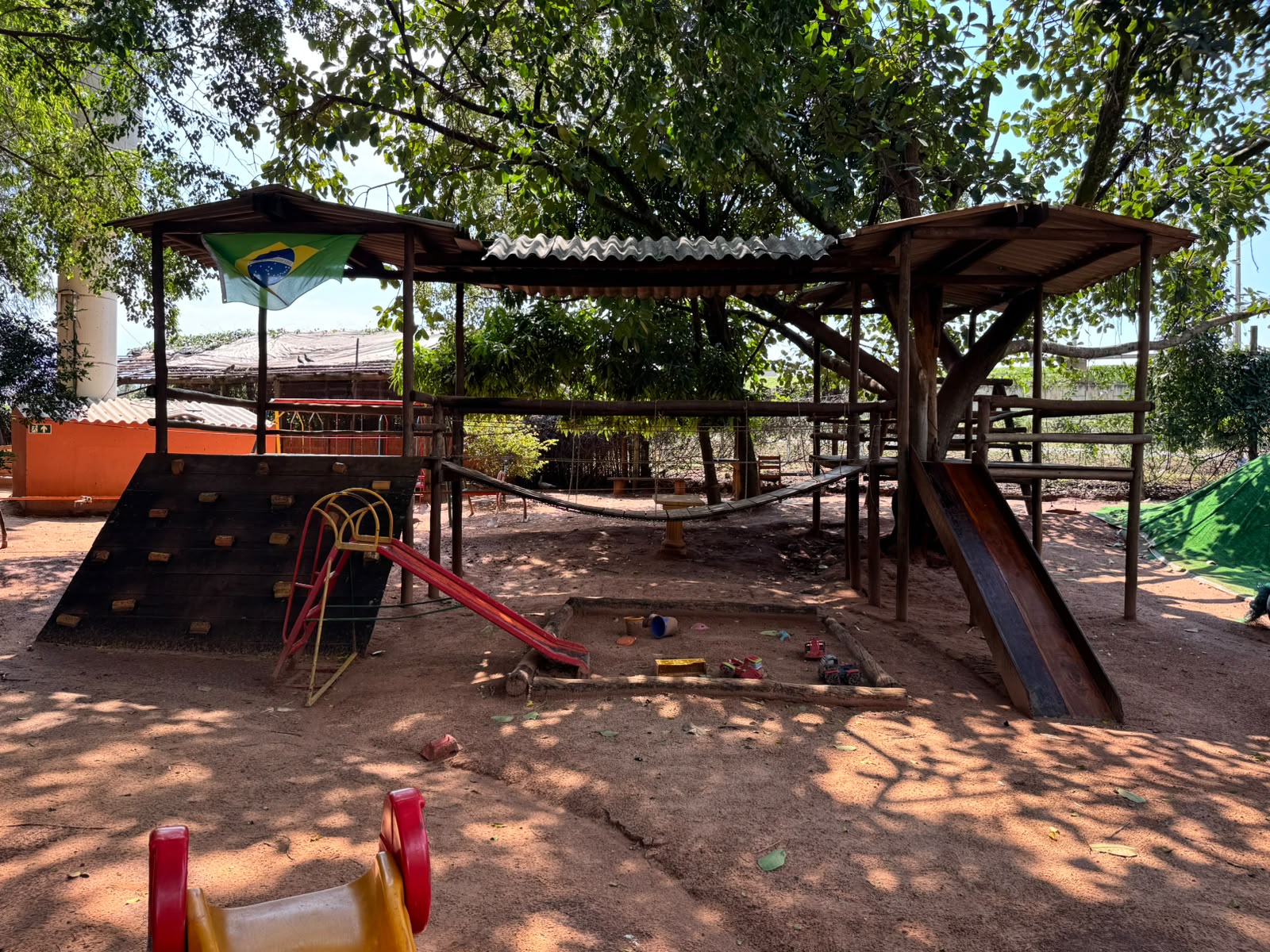 Playground de madeira construído ao redor de uma árvore, com ponte de cordas, caixa de areia e bandeira do Brasil