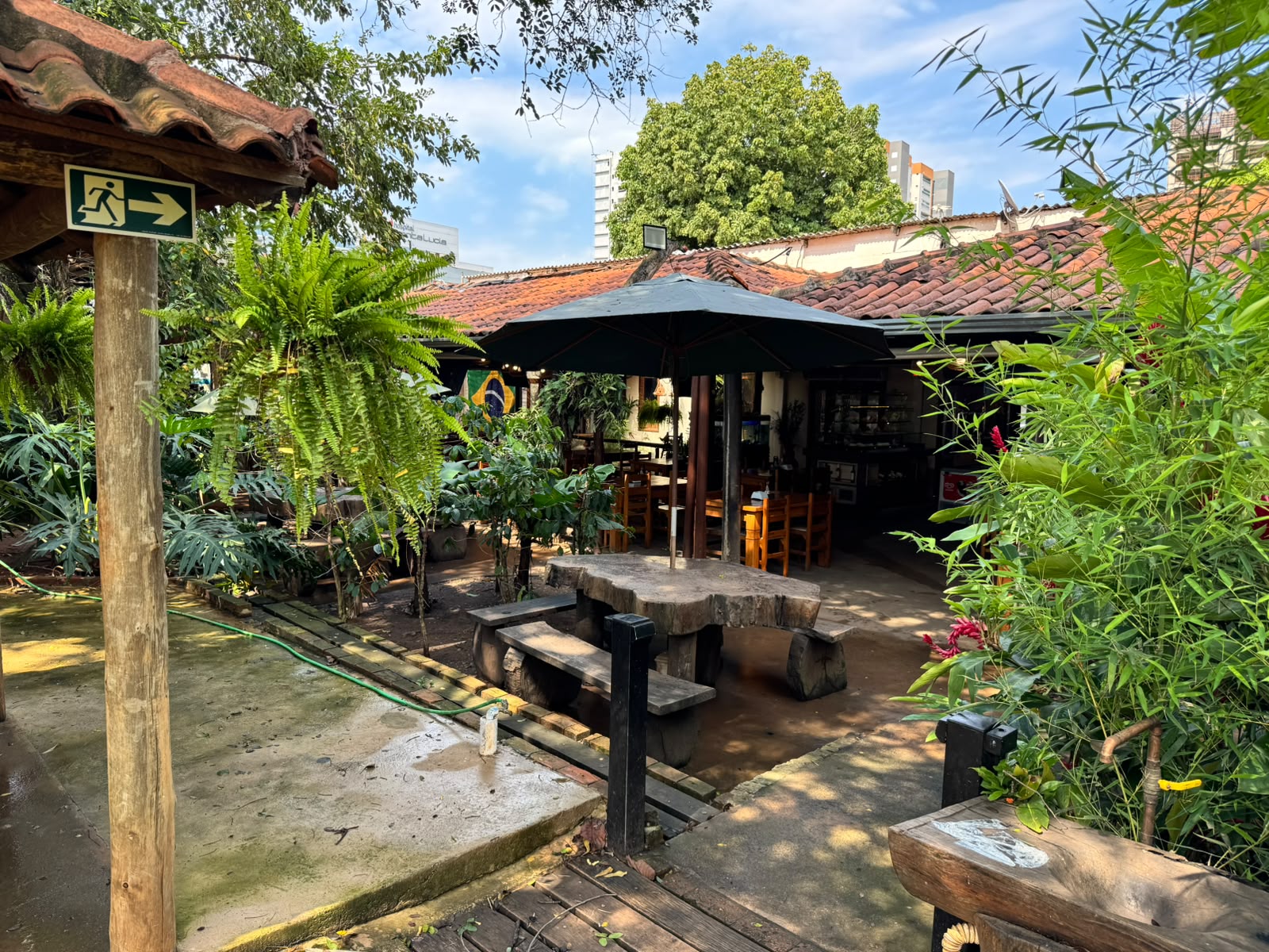 Mesa rústica de madeira com bancos sob ombrelone preto, cercada por jardins e mesas cobertas