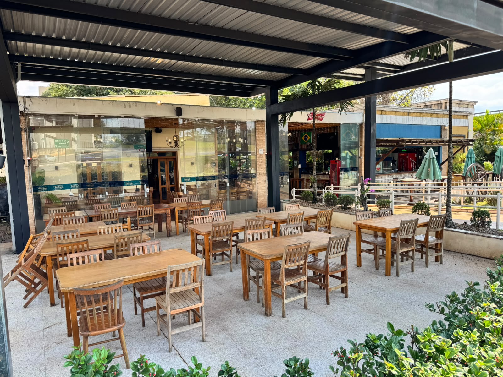 Pátio externo coberto com telhas metálicas, mesas de madeira e vista para o jardim